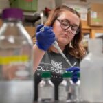 A woman with large round glasses is framed by lab equipment as she adds fluids into a test tube.