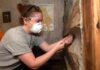 a student in a protective mask uses a wooden drill bit to extract a piece of wood from a cabin-like structure