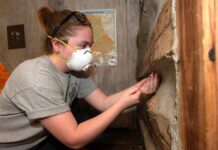 a student in a protective mask uses a wooden drill bit to extract a piece of wood from a cabin-like structure