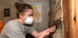 a student in a protective mask uses a wooden drill bit to extract a piece of wood from a cabin-like structure