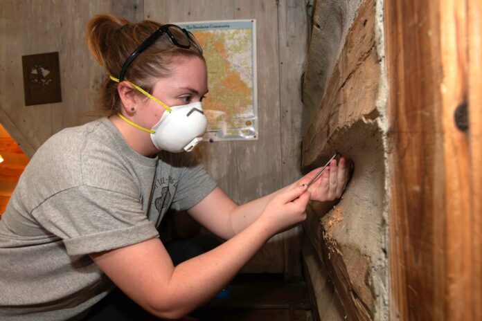 a student in a protective mask uses a wooden drill bit to extract a piece of wood from a cabin-like structure