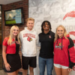 Four students--two men and two women, pose in front of a UofL banner.