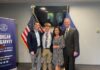 Congressman Morgan McGarvey, Jackson Robbins and Robbins' parents smile for a picture beside a United States flag and a Kentucky state flag.