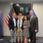 Congressman Morgan McGarvey, Jackson Robbins and Robbins' parents smile for a picture beside a United States flag and a Kentucky state flag.