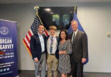 Congressman Morgan McGarvey, Jackson Robbins and Robbins' parents smile for a picture beside a United States flag and a Kentucky state flag.