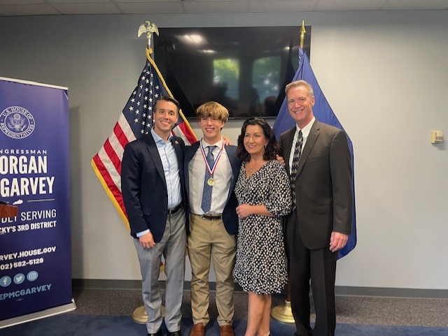 Congressman Morgan McGarvey, Jackson Robbins and Robbins' parents smile for a picture beside a United States flag and a Kentucky state flag.