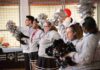 Five cheerleaders in a row, facing left, holding pom-poms. They are wearing grey sweatshirts and skirts or pants. They are standing in an indoor space with a basketball hoop and a banner with "Special Olympics Unified Champion Schools" visible in the background.