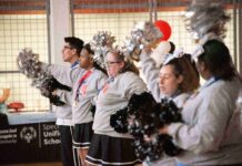 Five cheerleaders in a row, facing left, holding pom-poms. They are wearing grey sweatshirts and skirts or pants. They are standing in an indoor space with a basketball hoop and a banner with "Special Olympics Unified Champion Schools" visible in the background.