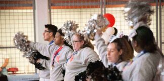 Five cheerleaders in a row, facing left, holding pom-poms. They are wearing grey sweatshirts and skirts or pants. They are standing in an indoor space with a basketball hoop and a banner with "Special Olympics Unified Champion Schools" visible in the background.