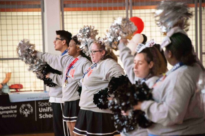 Five cheerleaders in a row, facing left, holding pom-poms. They are wearing grey sweatshirts and skirts or pants. They are standing in an indoor space with a basketball hoop and a banner with 